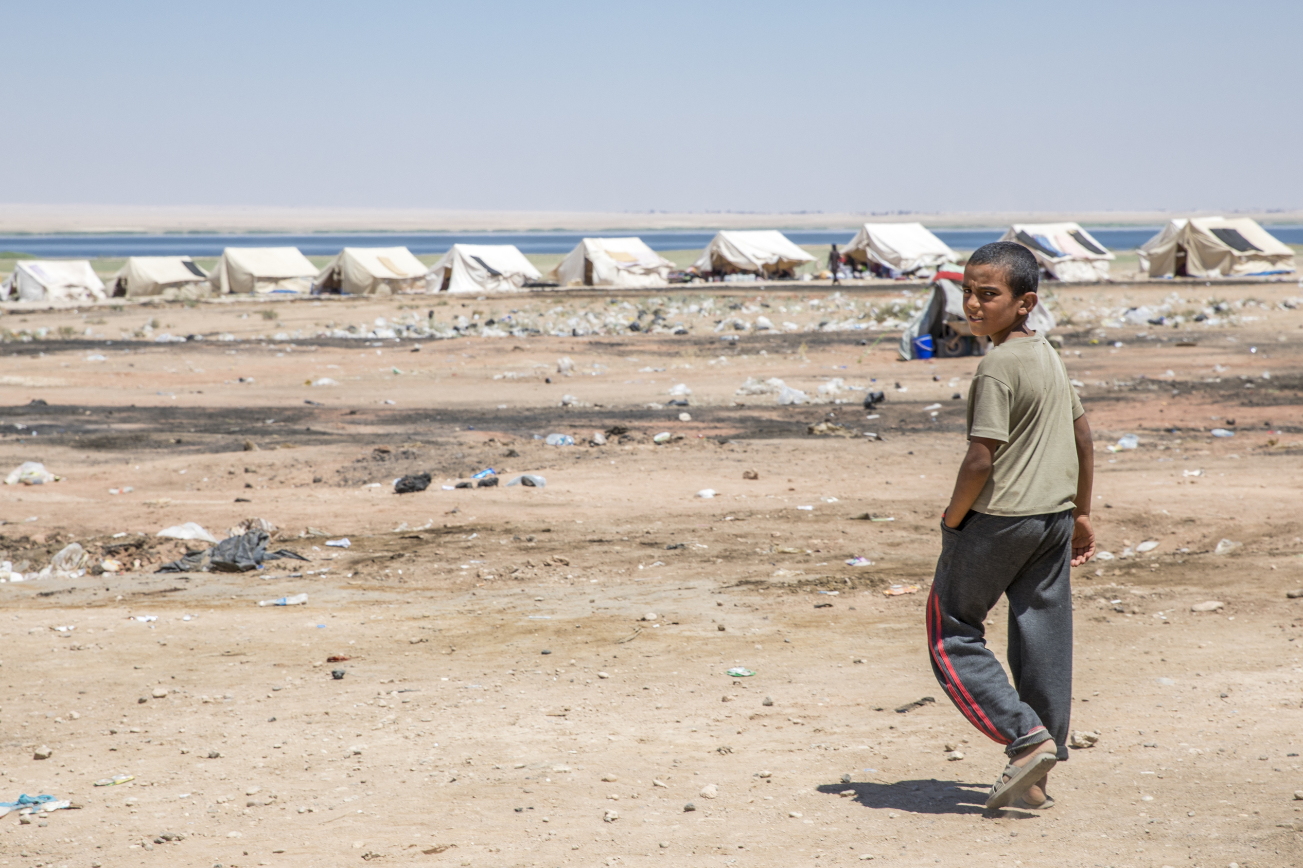 A boy walks toward a row of white tents in a dry, desert landscape