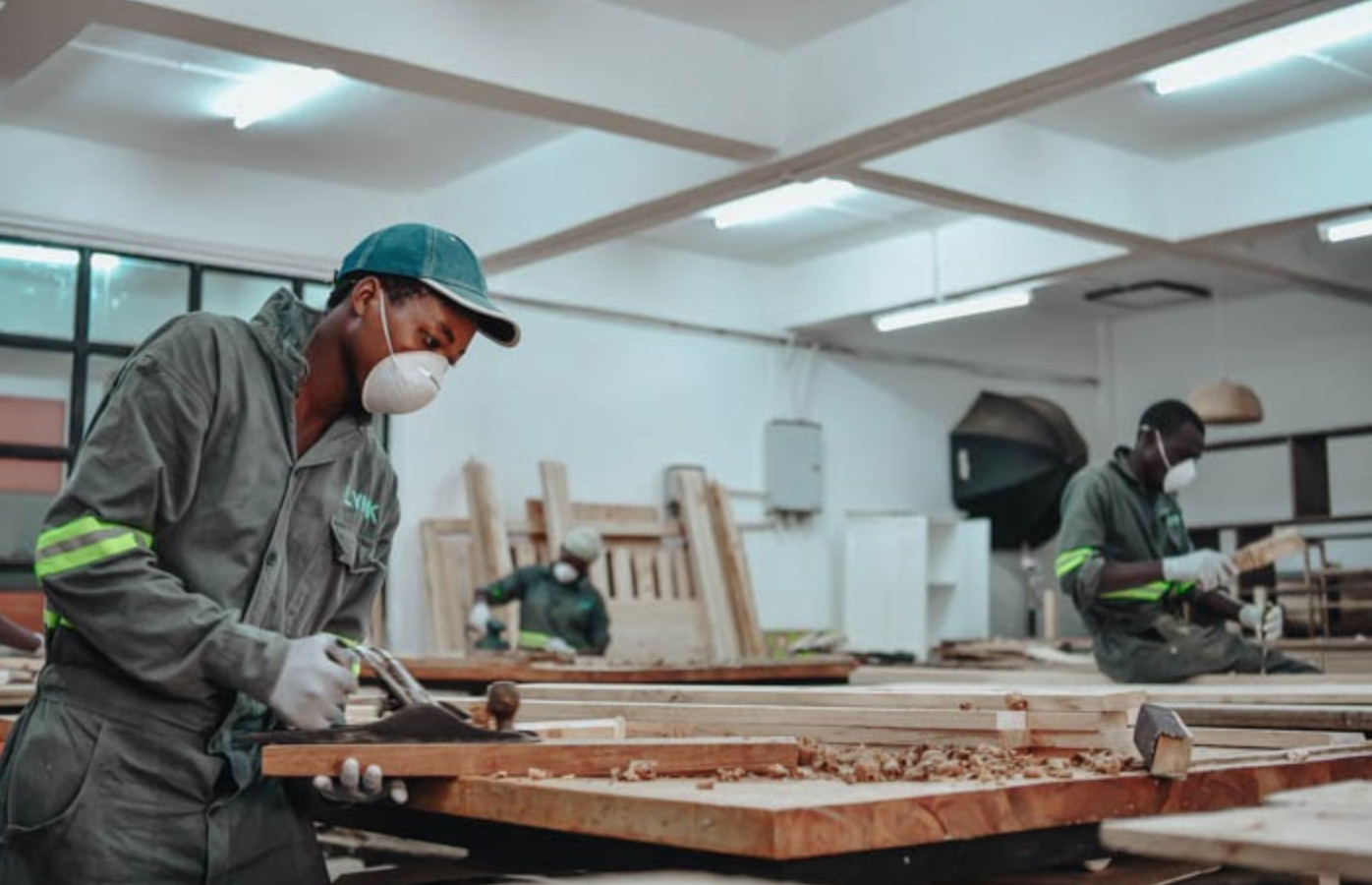 A person uses wood working tools while wearing a face mask in a room with other face masked workers.
