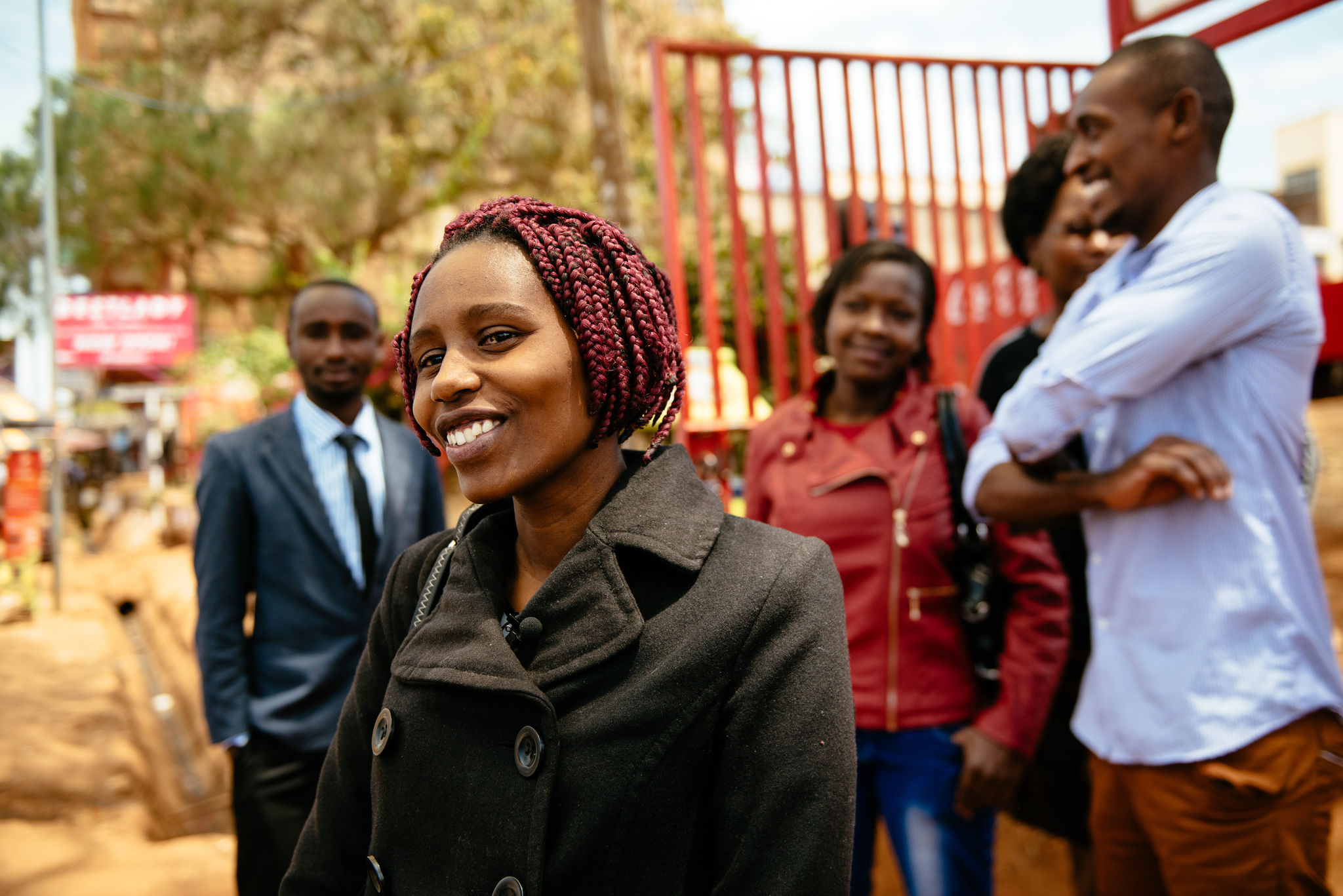 A smiling person in a black coat wears a lapel microphone while a group of smiling people stand behind her in Kenya.