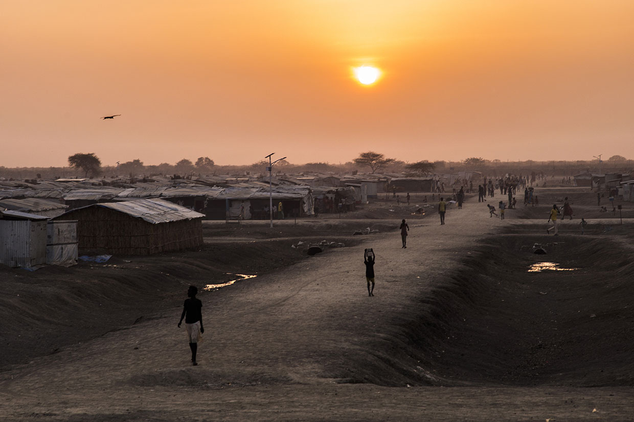 Individuals walking a road through a refugee camp at sunset.
