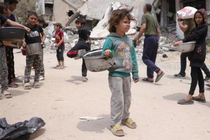 Palestinian children stand in a damaged public space with empty cooking pots.