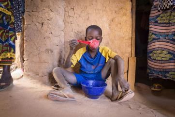 A boy in niger sitting against a wall eating from a blue bowl with a large red spoon