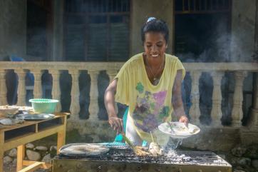 A woman in a yellow t-shirt prepares fish on a grill in timor leste
