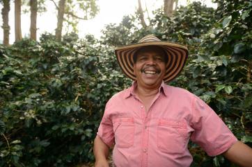 A person wearing a large brim hat smiles for the camera in a lush environment. 