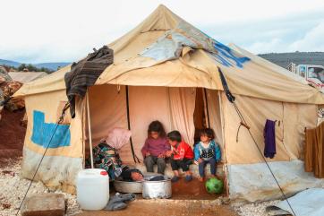 An adult washes clothes, while children sit at the door of their tent in kalli camp.