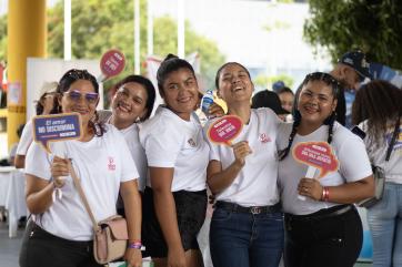 Group of woman stand among an event, holding signs and smiling.