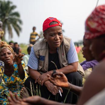 A Mercy Corps staff member crouches to speak to a group of people sitting down.