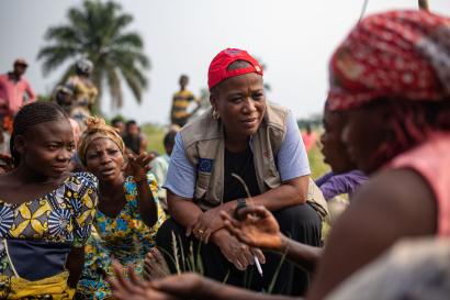 A mercy corps staff member crouches to speak to a group of people sitting down.