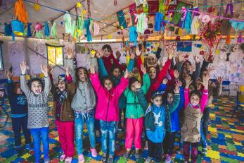 Group of child refugees smiling with their arms raised at a mercy corps youth center surrounded by bright decorations