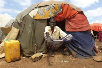 A man in a displacement camp in somalia sits in front of a tent that is covered in multiple different fabrics