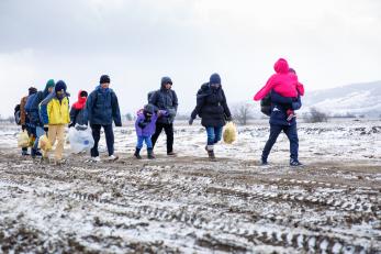Adults carry yellow plastic bags; others carry children as they walk across a snow-covered landscape