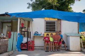 A man and three boys sit outside a home with a blue tarp roof