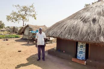 A person stands outside of a grass-thatched home 