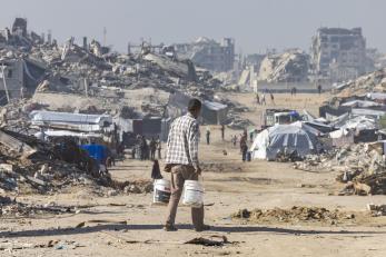 Palestinian man, holding buckets, walks among rubble of gaza.