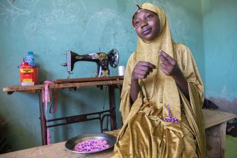 Nigerian woman beading, while seated, sewing machine in the background.