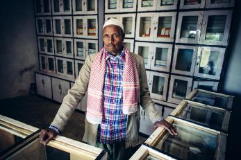 A man in ethiopia stands in a shop with supplies in cabinets behind him.