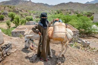 A person stands next to a livestock animal in yemen. the animal is carrying a plastic jug and other items on its back.