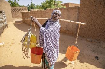 Halma, pictured in a purple and white shawl carrying three buckets balanced across her shoulders on a large stick