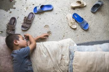 Iraqi child naps on the dirt floor inside tent, dusty sandals splayed beside him.