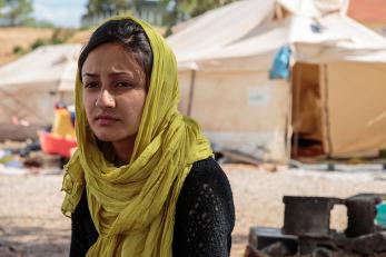 Saman, wearing a black top and green scarf, sitting in a refugee camp with a sad expression.