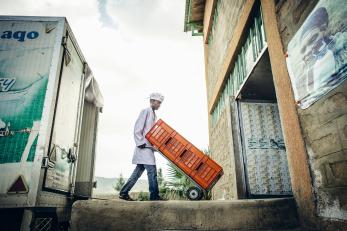 A man in a white coat and hat rolls orange crates from the milk truck into a building