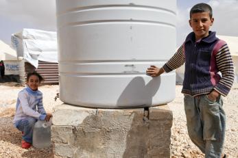 A girl and boy next to a large white water tank at a refugee camp in Jordan