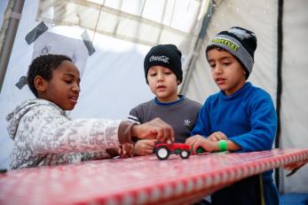 Three boys play with toy cars on a red and white tabletop