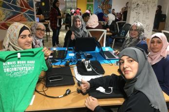 A group of young people sit a table with laptops and mobile devices and pose for the camera.