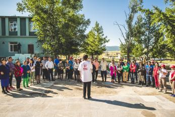 A person wearing a Mercy Corps shirt addresses a group of people arranged in a semicircle on a sunny day in Mongolia.