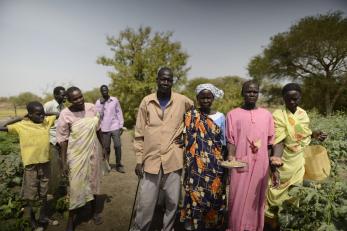 A group of people stand  outside together holding harvested agricultural goods in South Sudan. 