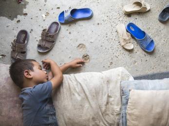 Iraqi child naps on the dirt floor inside tent, dusty sandals splayed beside him.