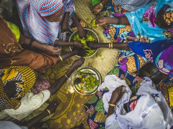 Women in colorful clothing reaching in to share food from a bowl at center in niger. photo: sean sheridan for mercy corps