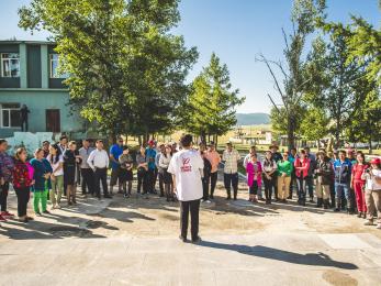 A person wearing a mercy corps shirt addresses a group of people arranged in a semicircle on a sunny day in mongolia.
