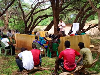 Kenyan community members meet in the outdoors under trees.