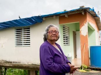 A woman with gray hair and a purple cardigan stands outside a home in puerto rico